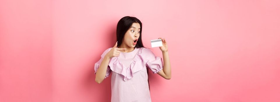 Excited asian woman pointing and showing plastic credit card, gasping amazed, concept of shopping.