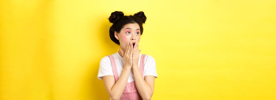 Shocked teen asian girl with makeup, gasping and covering mouth, looking left at logo amazed, standing on yellow background.