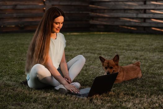 Lifestyle girl and Corgi dog outside. Girl with laptop petting her Welsh Corgi Pembroke dog on the grass. Busy young woman have a break and relax with her dog. Lifestyle with domestic pet