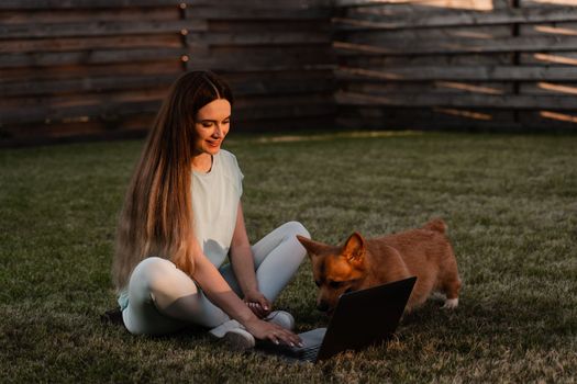 Lifestyle girl and Corgi dog outside. Girl with laptop petting her Welsh Corgi Pembroke dog on the grass. Busy young woman have a break and relax with her dog. Lifestyle with domestic pet