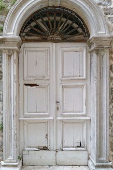 The impressive double-leaf entrance gate of a manor house is decorated with a round arch with window and grille. The white color is already peeling off in some places.