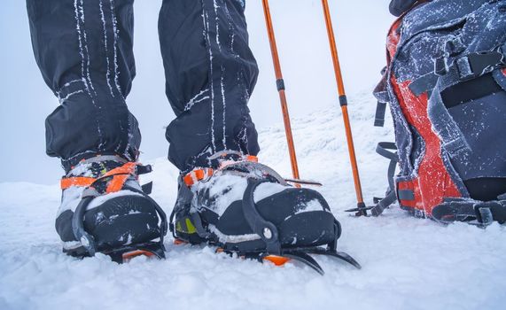 Man wearing crampons standing beside backpack and trekking sticks on deep snow