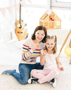 Cute little girl and mother holding stick glasses in kids room