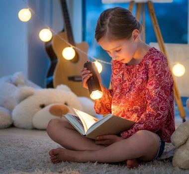 Cute little girl reading book at night using flashlight indoors