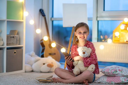 Beautiful girl with smartphone sitting on the floor in the bedroom. Child with phone and Teddy bear smiling. Bedtime with magical lights