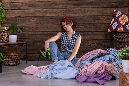 A young girl is waiting to iron men's shirts. She sits on the floor of a room in the house. An overturned basket of clothes. A wall of brown boards.