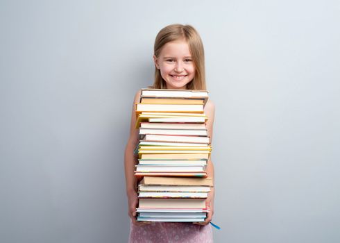 Primary school girl holding stack of books on a grey wall background