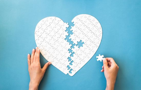 White puzzle in heart shape. Hands connect white details of puzzle on blue background.