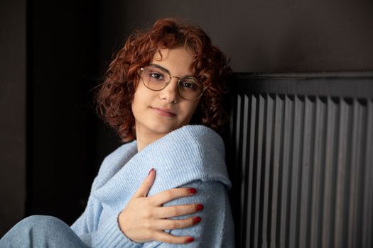 A girl in a blue thick sweater sits by the radiator. Poorly working central heating. A gentle smile.