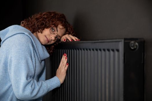 A young girl in a blue thick sweater hugs a warm radiator. Central heating necessary in the house during the winter period.