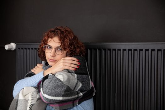 A girl with an imposed scarf leans with her back against a radiator. An attractive woman is freezing in her own home due to a central heating malfunction.