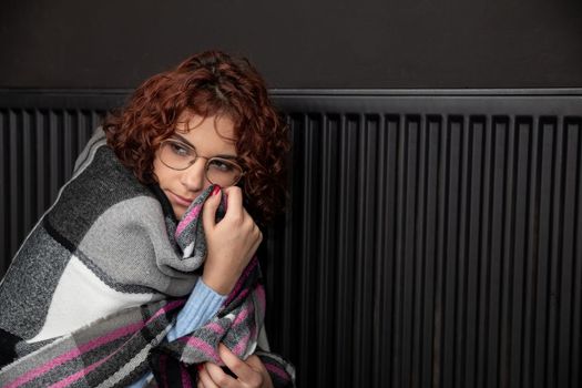 A girl sits wrapped in a scarf hoping the heater will finally start heating. Central heating during the first startup.