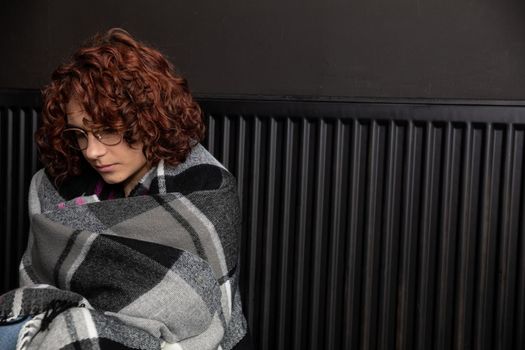 A woman sits on the floor with her back leaning against a radiator. The adult girl has a thick checkered scarf on her back. Central heating.