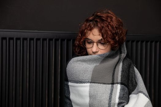 A woman sits on the floor with her back leaning against a radiator. The adult girl has a thick checkered scarf on her back. Central heating.