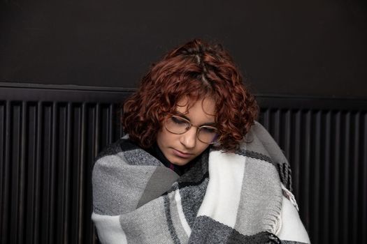 A girl with an imposed scarf leans with her back against a radiator. An attractive woman is freezing in her own home due to a central heating malfunction.