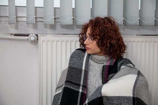 A woman sits on the floor with her back leaning against a radiator. The adult girl has a thick checkered scarf on her back. Central heating.