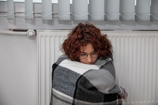 A girl with an imposed scarf leans with her back against a radiator. An attractive woman is freezing in her own home due to a central heating malfunction.