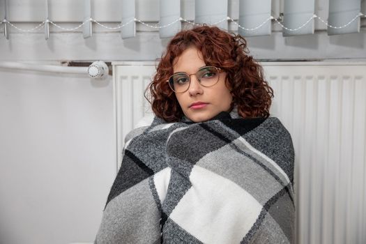 A girl with an imposed scarf leans with her back against a radiator. An attractive woman is freezing in her own home due to a central heating malfunction.