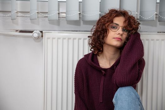 Sad woman sitting by the radiator. Faulty central heating. It is time for the heating season in apartment blocks.