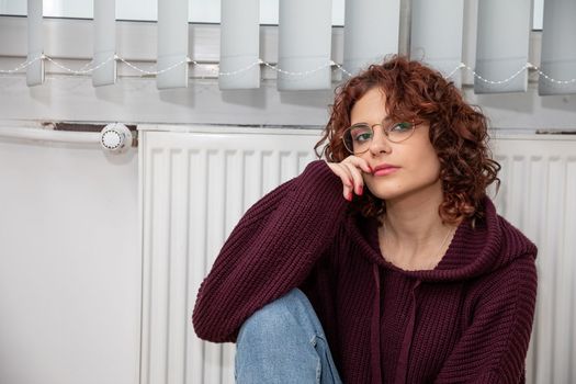 Dressed in a thick sweater, the girl sits on the floor by the radiator hoping that they will finally start the central heating.