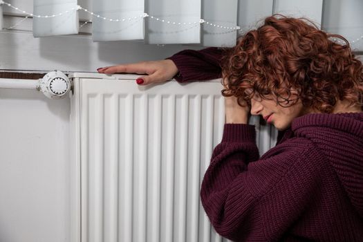 Dressed in a thick sweater, the girl sits on the floor by the radiator hoping that they will finally start the central heating.