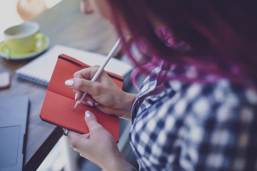 Positive female student with pink hair writing down funny story in notebook sitting in coffee shop with cup of cappuccino and laptop. Smiling female noting text in notepad