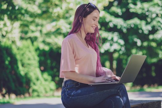 Young girl with pink hair is studying in the spring park, sitting on the wooden bench and browsing on her laptop