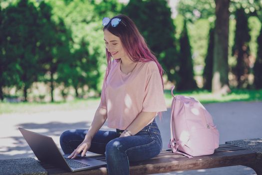 Young girl with pink hair is studying in the spring park, sitting on the wooden bench and browsing on her laptop