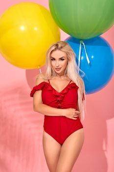 Young sexy slim woman in a red swimsuit with balloons in her hand hugged herself. Full length fashion portrait of a beautiful girl with long blond hair, on a pink background.