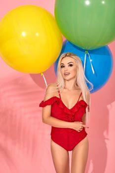 Sexy slim woman in a red swimsuit with balloons in her hand hugged herself. Full length fashion portrait of a beautiful girl with long blond hair, on a pink background.