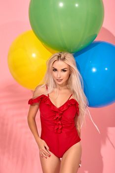Charming slim woman in a red swimsuit with balloons in her hand is posing in a studio. Full length fashion portrait of a beautiful girl with long blond hair, on a pink background.