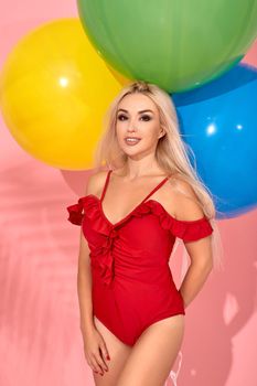 Wonderful slim woman in a red swimsuit with balloons in her hand is posing sideways in a studio. Full length fashion portrait of a beautiful girl with long blond hair, on a pink background.