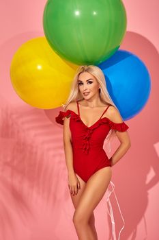 Stunning blonde female model with amazing body standing in an elegant red swimsuit on pink background with palm shadow. Vacation and recreation concept