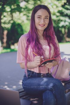 Woman writing in a notebook sitting on a wooden bench in the park. Girl working outdoors on portable computer, copy space. Technology, communication, freelance and remote working concept.