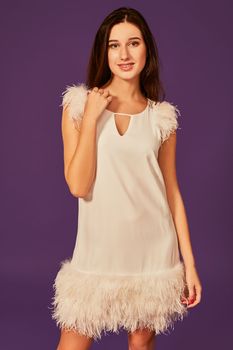 Beautiful brunette woman in elegant cocktail white dress is posing in studio on purple background. Three quarter length studio shot. Holding hand on her neck