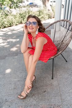 Portrait of a woman with glasses. Photo of a fashionable girl with beautiful brown hair, dressed in a red sundress with polka dots, sits in a chair on the street