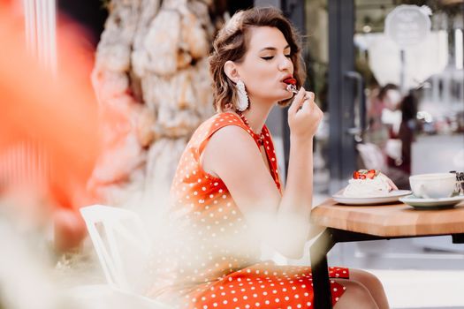 Charming woman in a restaurant, cafe on the street. She sits at the table and eats a cake with a fork. Dressed in a red sundress with white polka dots