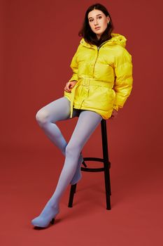 Stylish photo of cute brunette girl wearing yellow oversized down jacket posing in studio on red background, sitting on bar stool