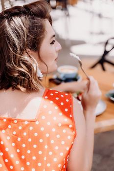 Charming woman in a restaurant, cafe on the street. She sits at the table and eats a cake with a fork. Dressed in a red sundress with white polka dots