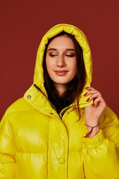 Close up photo of cute girl with dark hair in studio, in bright yellow down jacket. Red background, oversized clothes, hood on