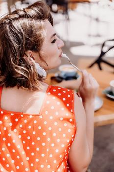 Charming woman in a restaurant, cafe on the street. She sits at the table and eats a cake with a fork. Dressed in a red sundress with white polka dots