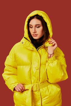 Close up photo of cute girl with dark hair in studio, in bright yellow down jacket. Red background, oversized clothes, hood on