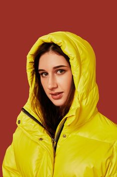 Close up photo of cute girl with dark hair in studio, in bright yellow down jacket. Red background, oversized clothes, hood on