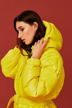 Close up photo of cute girl with dark hair in studio, in bright yellow down jacket. Red background, oversized clothes, hood off
