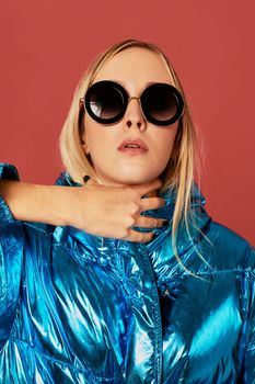 Concept art photo shot of slim blonde girl in studio, wearing blue down jacket, sunglasses, posing on camera, holding her neck.