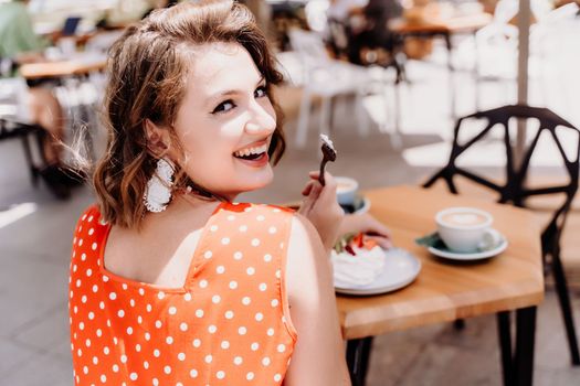 Charming woman in a restaurant, cafe on the street. She sits at the table and eats a cake with a fork. Dressed in a red sundress with white polka dots