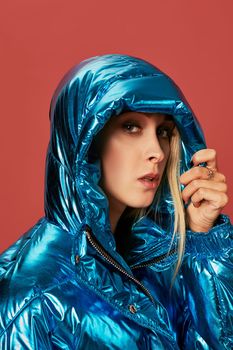 Close up fashion photo of young woman covering herself with hood. She wears blue oversized down jacket in studio on red background