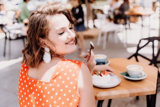 Charming woman in a restaurant, cafe on the street. She sits at the table and eats a cake with a fork. Dressed in a red sundress with white polka dots