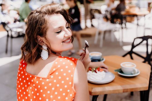 Charming woman in a restaurant, cafe on the street. She sits at the table and eats a cake with a fork. Dressed in a red sundress with white polka dots