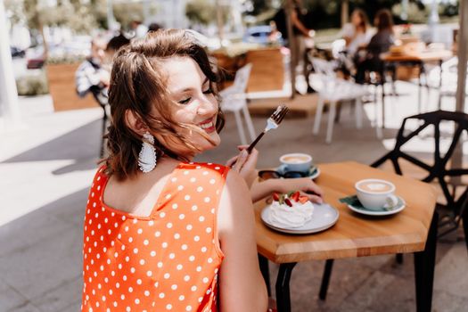 Charming woman in a restaurant, cafe on the street. She sits at the table and eats a cake with a fork. Dressed in a red sundress with white polka dots